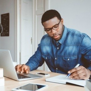 A man in a denim shirt is using a laptop and writing in a notebook.