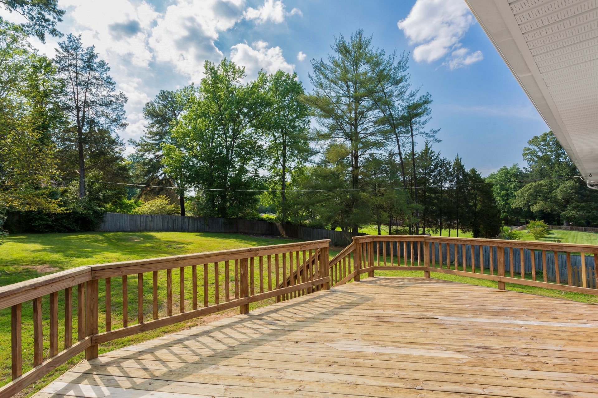 Wooden deck overlooking a grassy yard, with trees and a fence in the background under a bright blue sky.