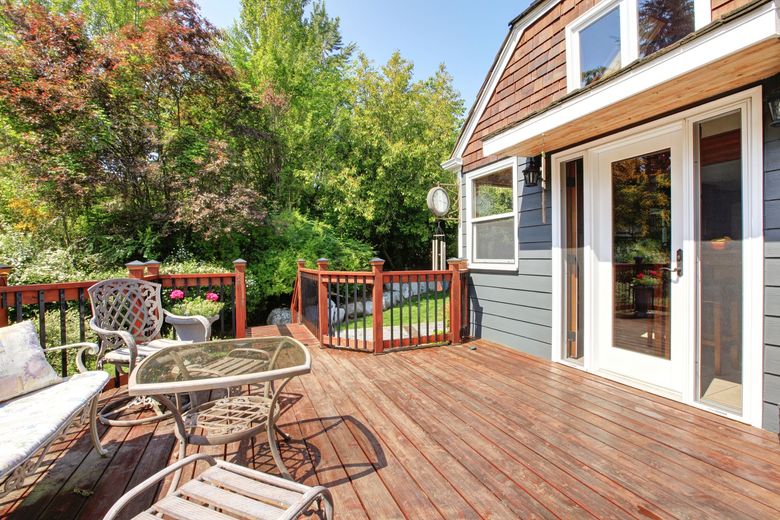 Wooden deck with furniture next to a house with a door, surrounded by trees.