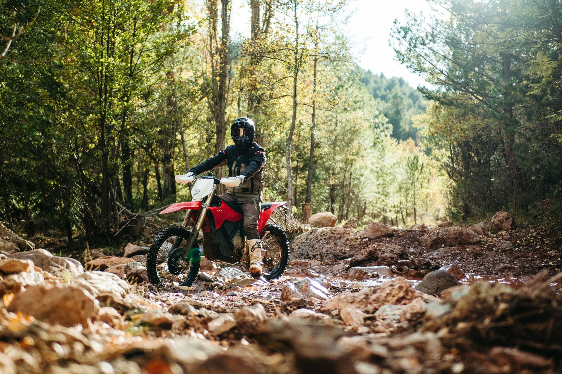 Person on a red dirt bike riding through a rocky stream in a forest.— EVA Motors in Paget, QLD