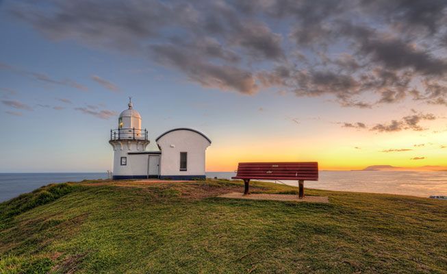 A Lighthouse and a Bench on Top of a Grassy Hill Overlooking the Ocean at Sunset — Hastings Garage Door Services in Port Macquarie
