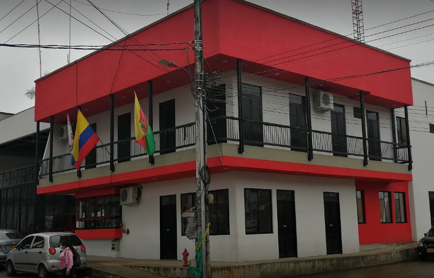 Un edificio blanco de dos plantas con una fachada superior de color rojo brillante, balcones en las esquinas y la bandera colombiana ondeando en la fachada principal.