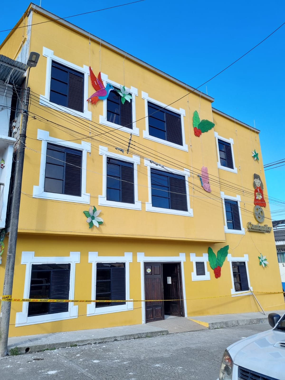 Un edificio de tres plantas de color amarillo brillante, con coloridas decoraciones florales y de colibríes en las paredes, y una entrada a pie de calle.