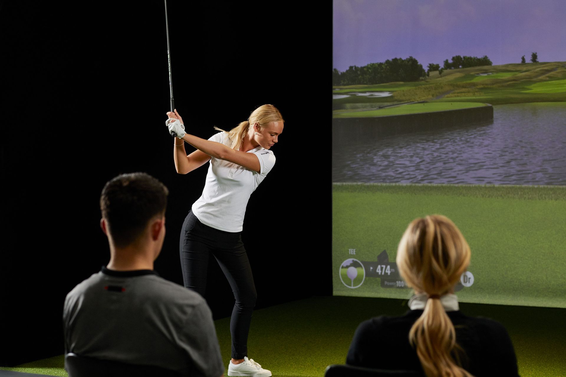 Woman swinging a golf club indoors, viewed by two observers, with a golf course displayed on a screen behind them.