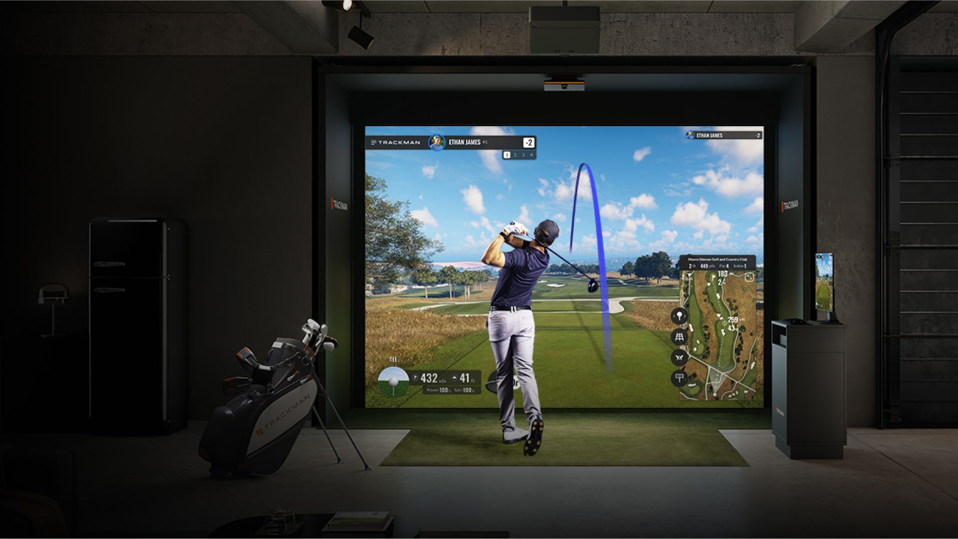 A golfer swinging in an indoor golf simulator; screen displays course; black and white, indoors.
