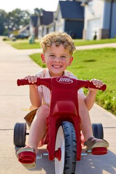 Boy with curly blonde hair smiles while riding a red tricycle on a sidewalk.