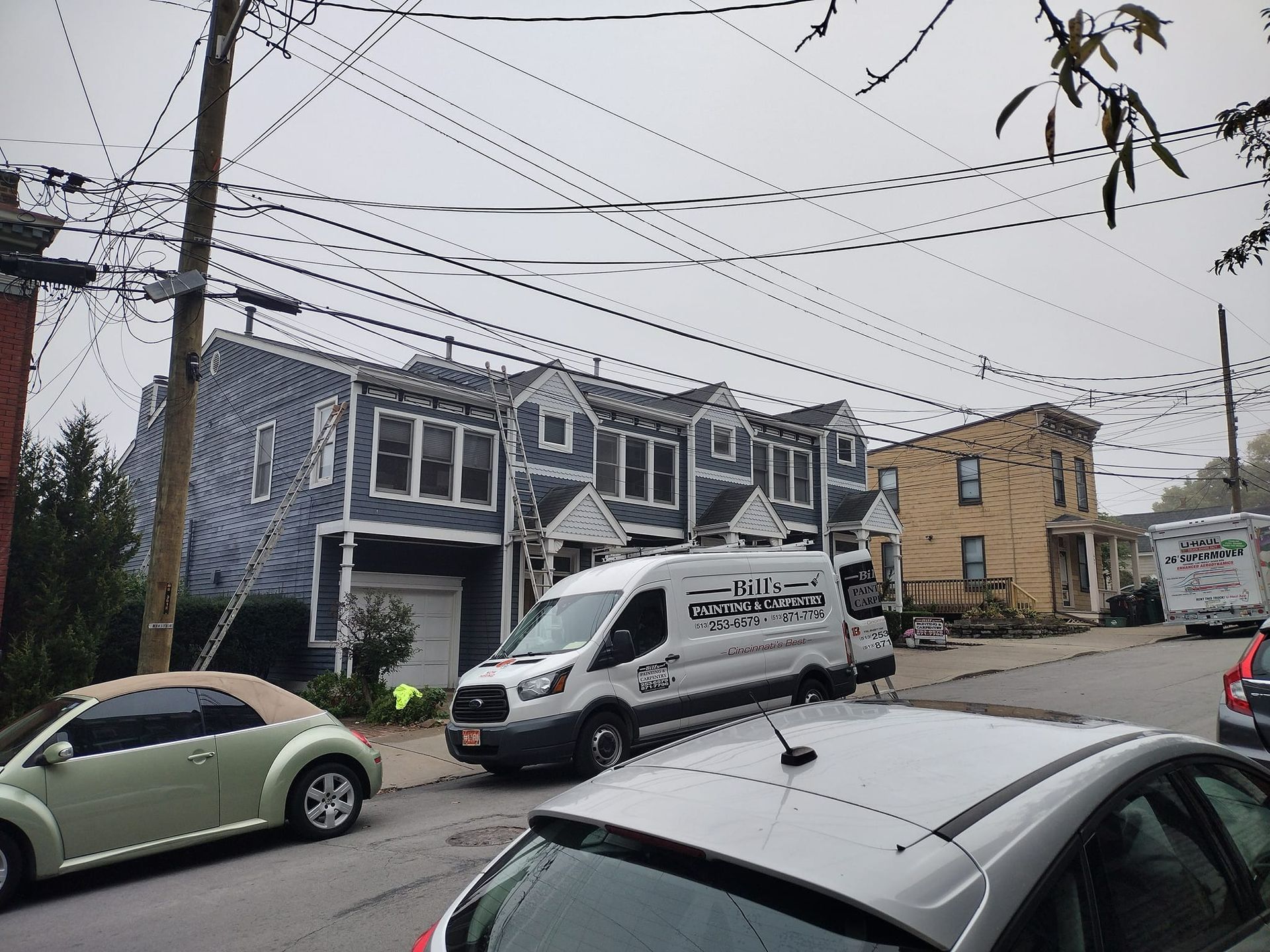 A city street scene with townhouses, power lines, vehicles, and a utility van on an overcast day.