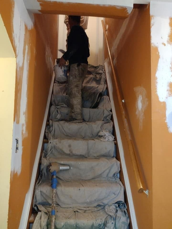 Person painting orange walls of a staircase. Drop cloths cover steps, with a paint can and paint brush.