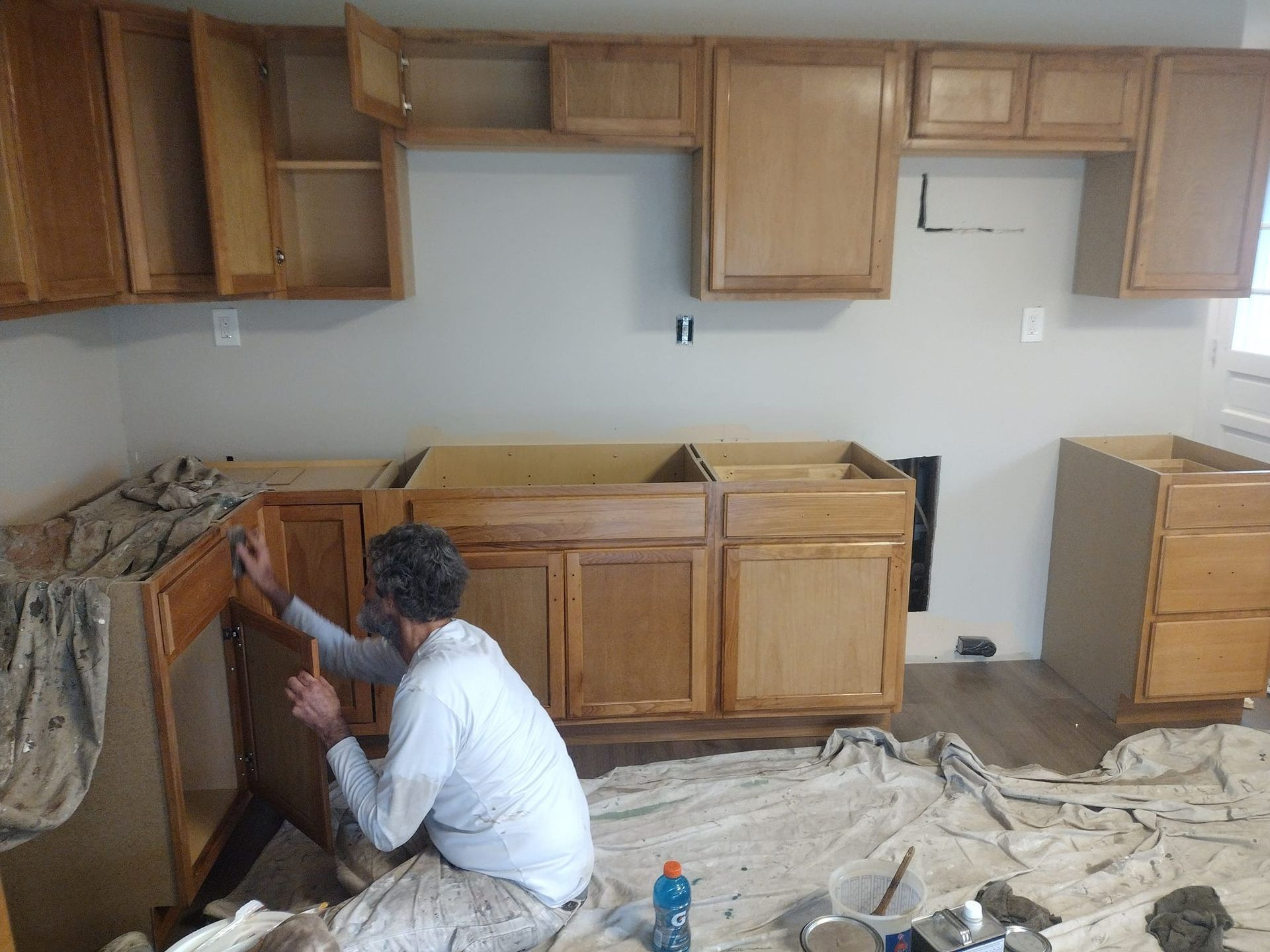Person painting kitchen cabinets in a room. Cabinets are light brown; walls are gray.