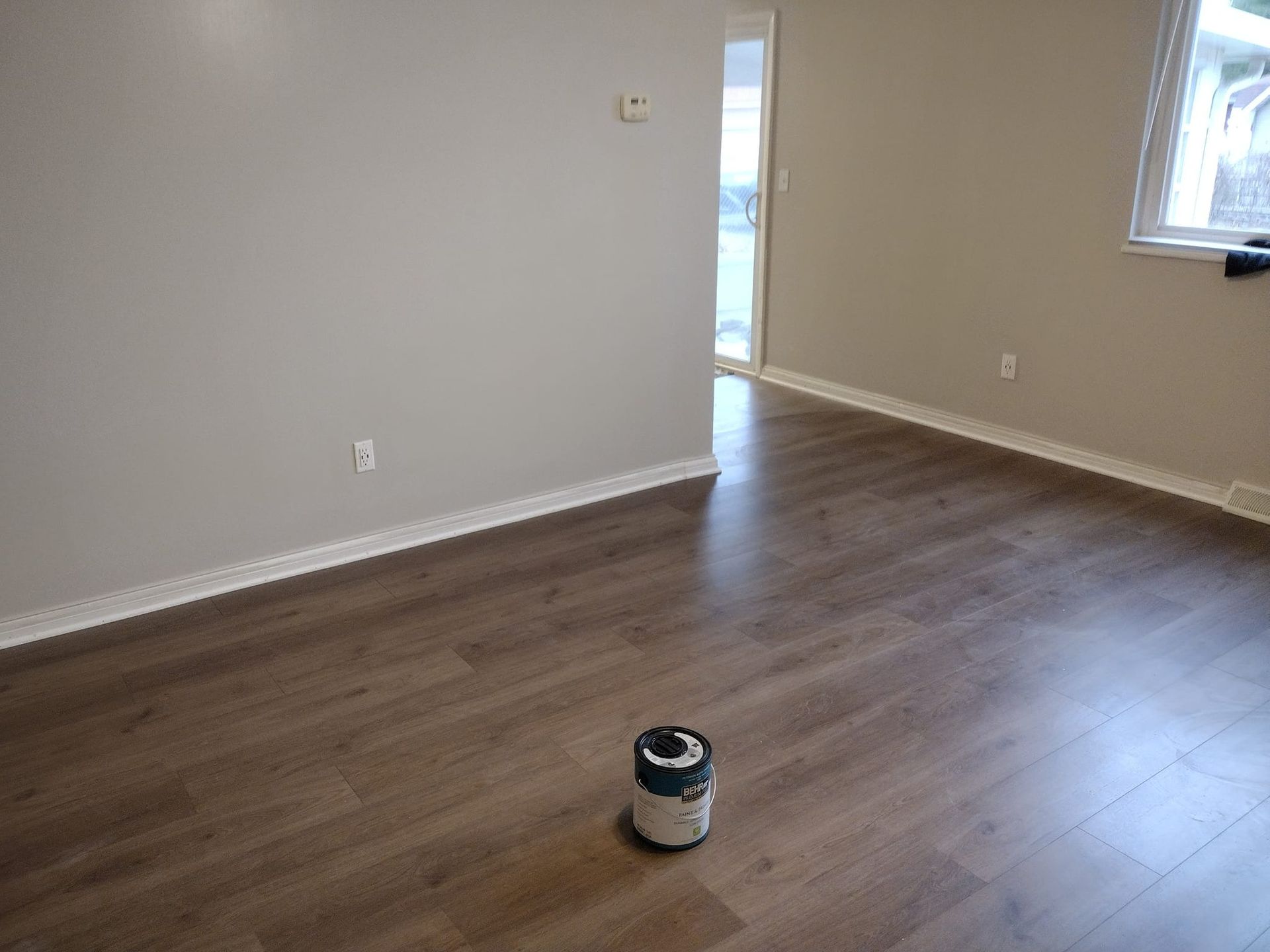 Empty room with gray walls, brown flooring, white trim, and a paint can on the floor.