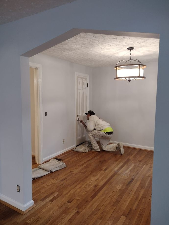 Person in protective suit painting a room with hardwood floors, arched doorway.