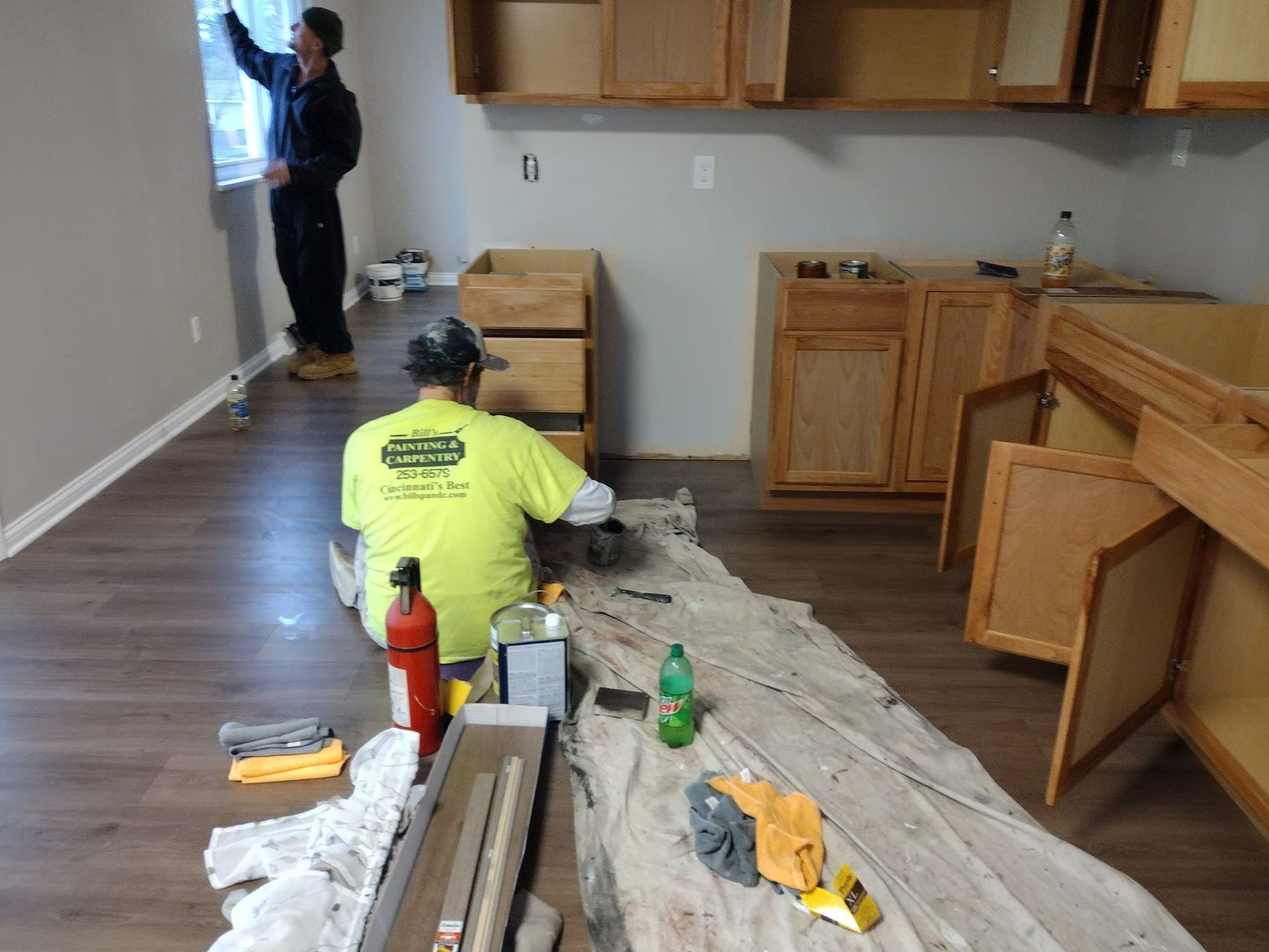 Two workers painting a kitchen. One paints a wall, the other works on a cabinet. Unfinished wood cabinets in a room.