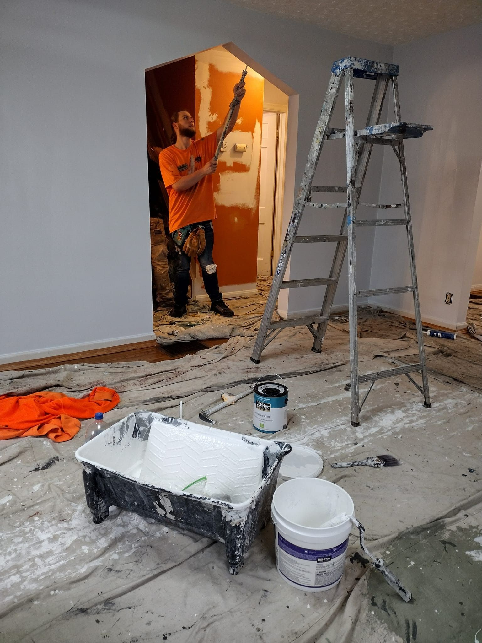 Man painting walls in a room. Gray walls, white paint supplies, ladder, orange shirt, messy drop cloth.