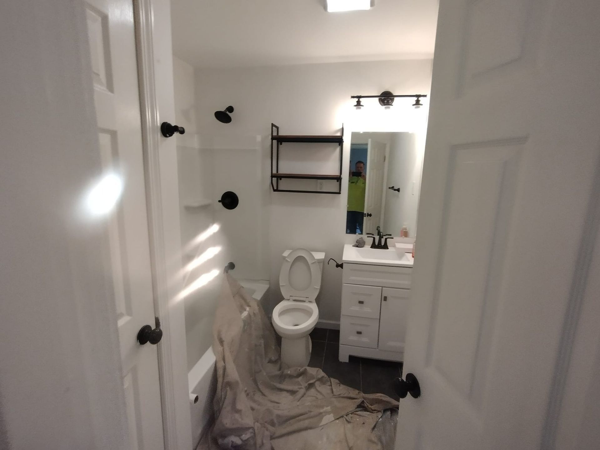 White bathroom with black fixtures, toilet, sink, and storage shelf; door open.