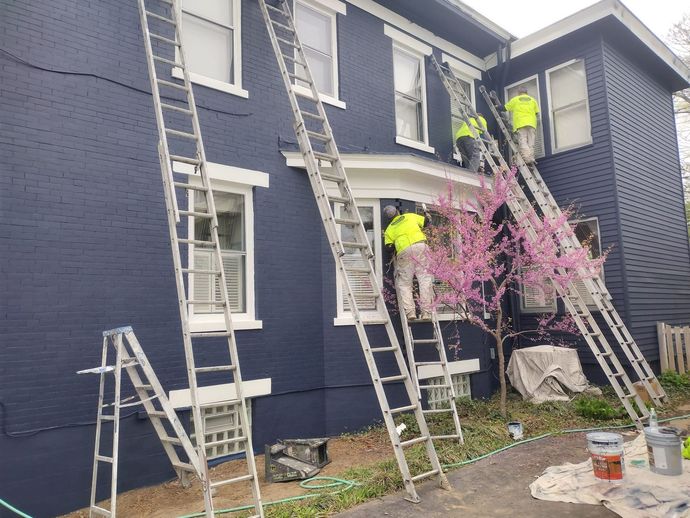 Workers on ladders painting a dark blue house exterior; bright yellow safety vests.
