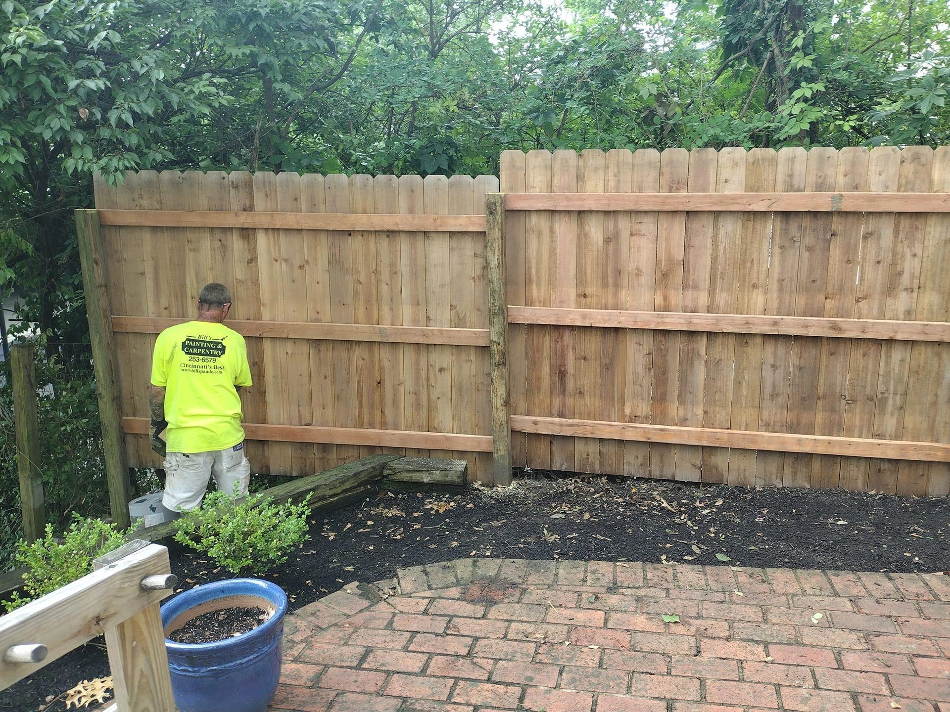 Person in yellow shirt repairing a wooden fence, in a backyard with a brick patio.