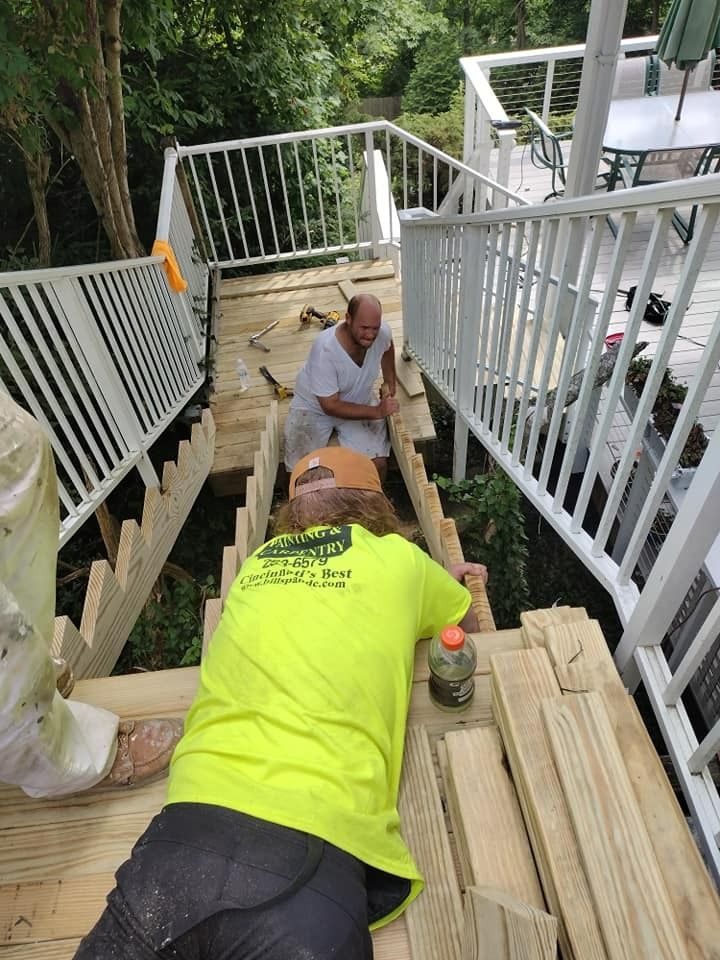Two men building wooden stairs outdoors, surrounded by white railings and trees. One wears neon shirt.
