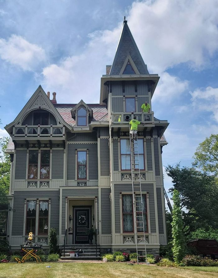 Victorian house with workers on a ladder. Gray exterior with beige trim, dark roof, and a tower.