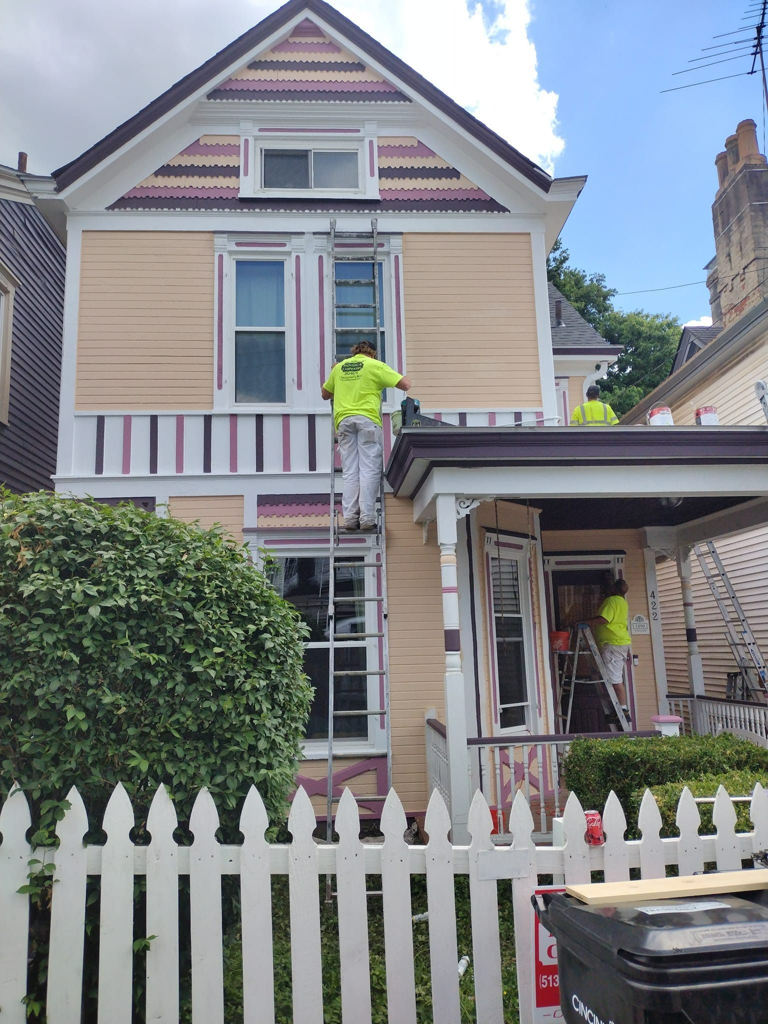 House exterior being painted by two workers in neon vests. White picket fence in the foreground.