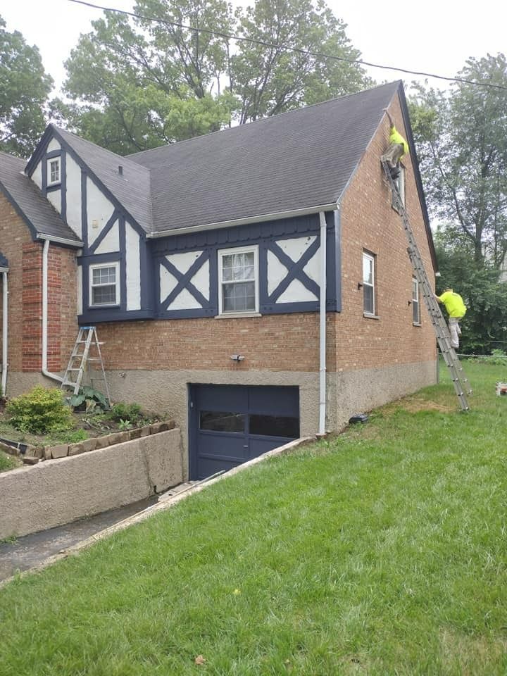 Two people on ladders working on the side of a brick and Tudor-style house.