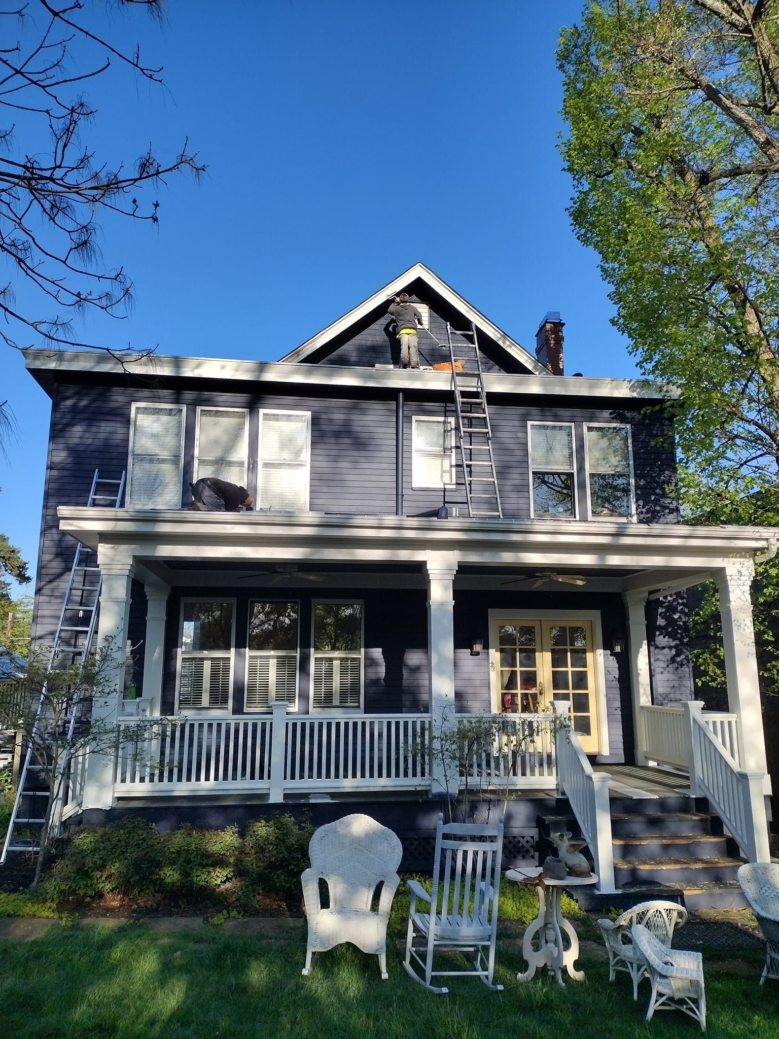 Two-story blue house with white porch, a ladder on the side, and several rocking chairs on the lawn.