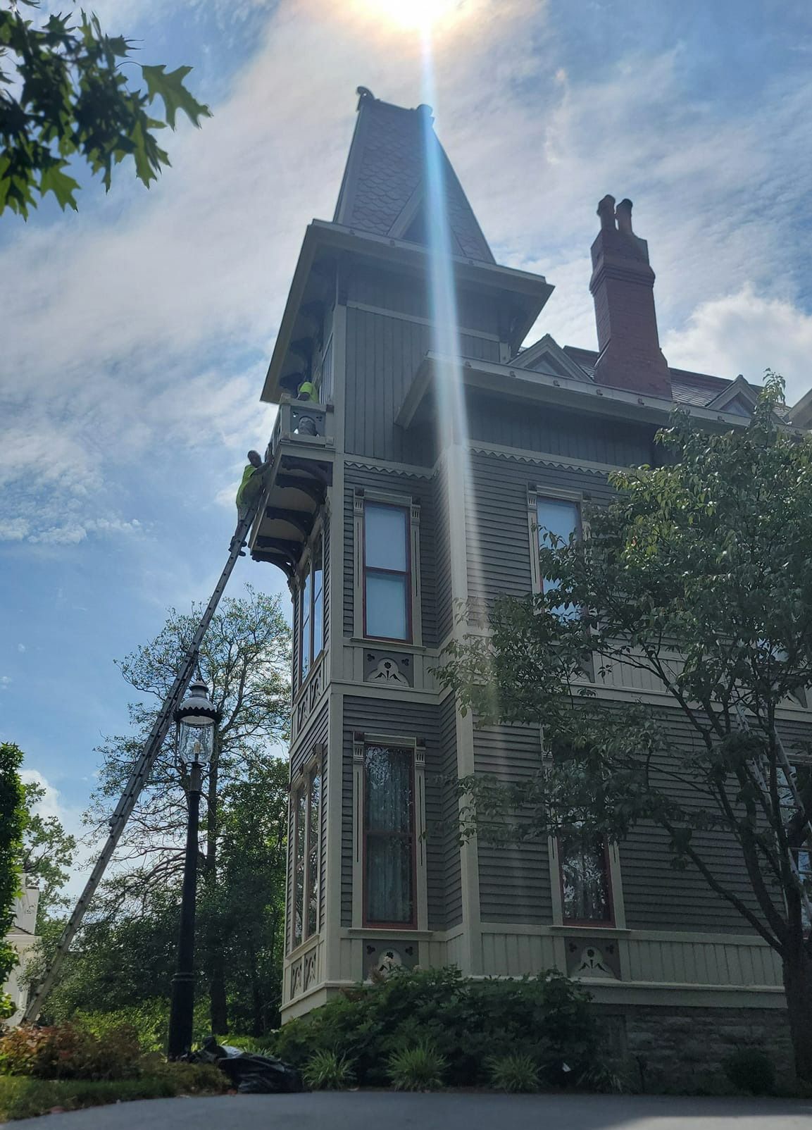Victorian house under bright sunlight. Men on lift working on the ornate roof, near tall trees.