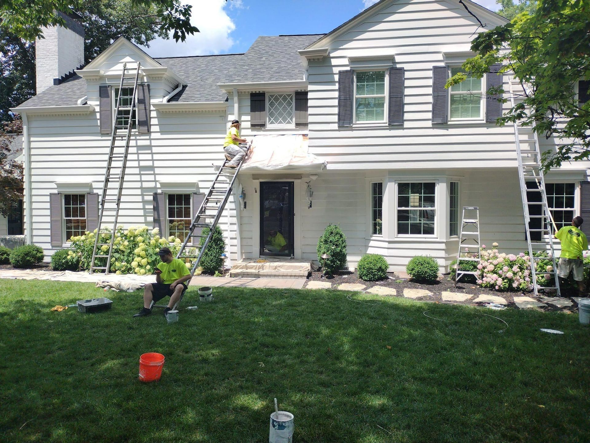 House being painted white by three workers on ladders; green lawn, blue sky.