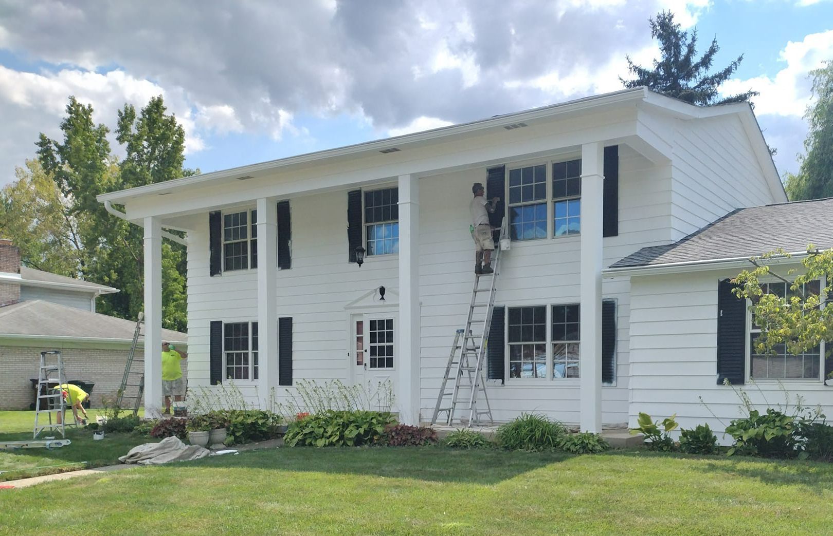 White two-story house with black shutters and columns; workers on a ladder; green lawn and cloudy sky.