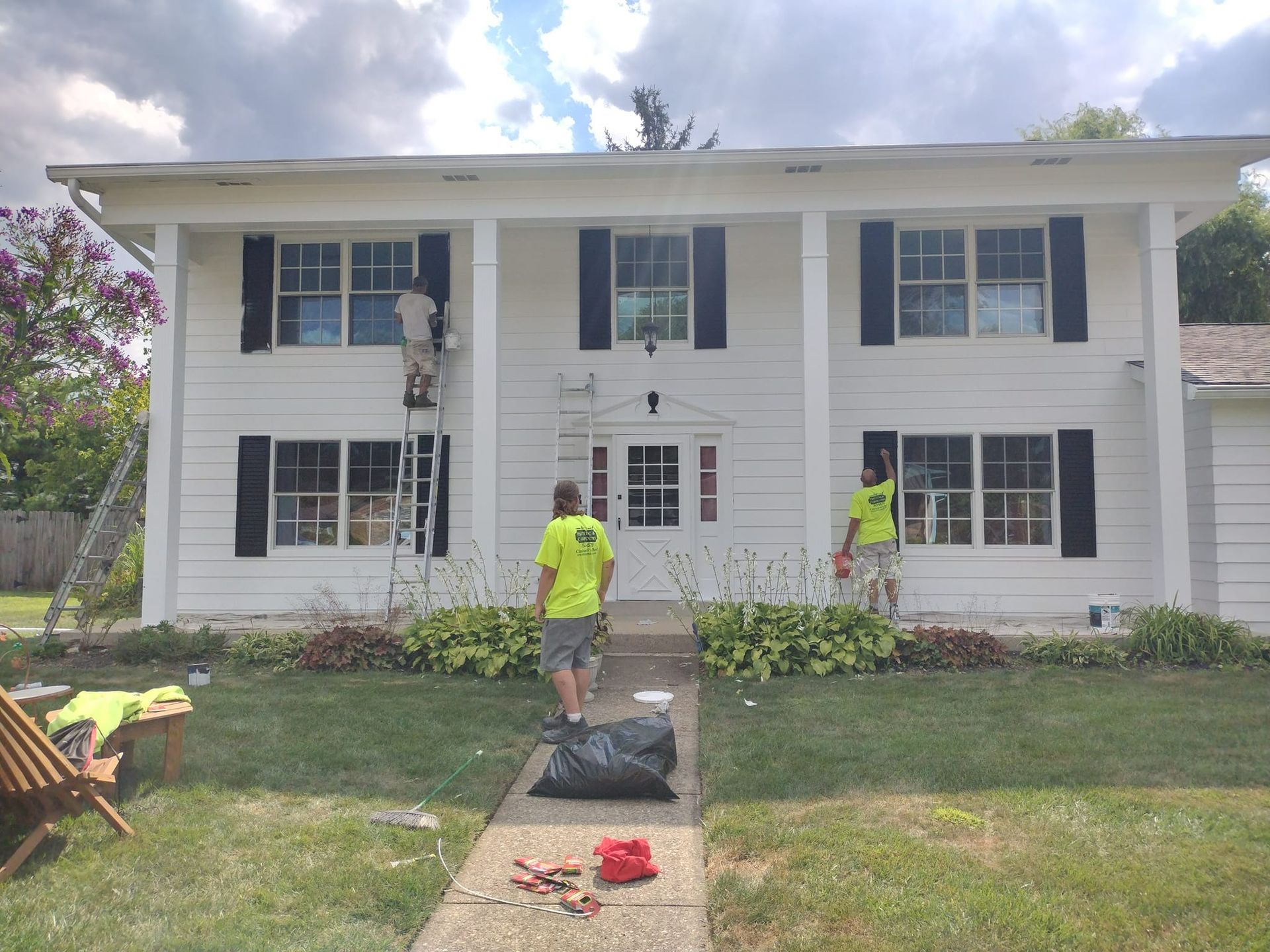 Workers painting a white two-story house with black shutters; one on a ladder, two others on the ground in the yard.