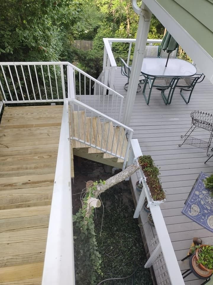Deck with stairs, white railings, gray and wood-colored surfaces. Garden below. Green plants and trees surround it.