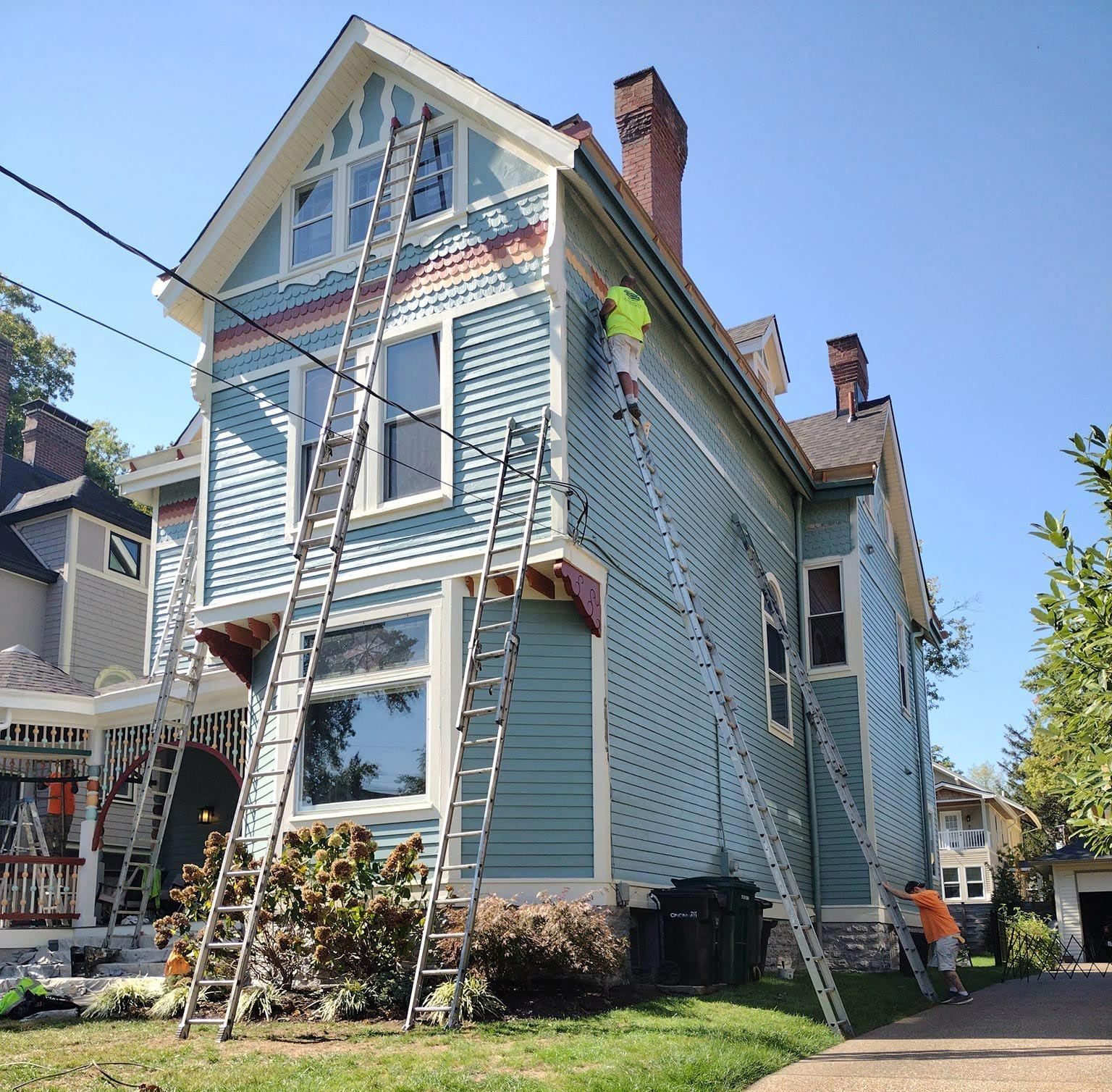 People painting a two-story Victorian house, light blue siding, multiple ladders, sunny day.