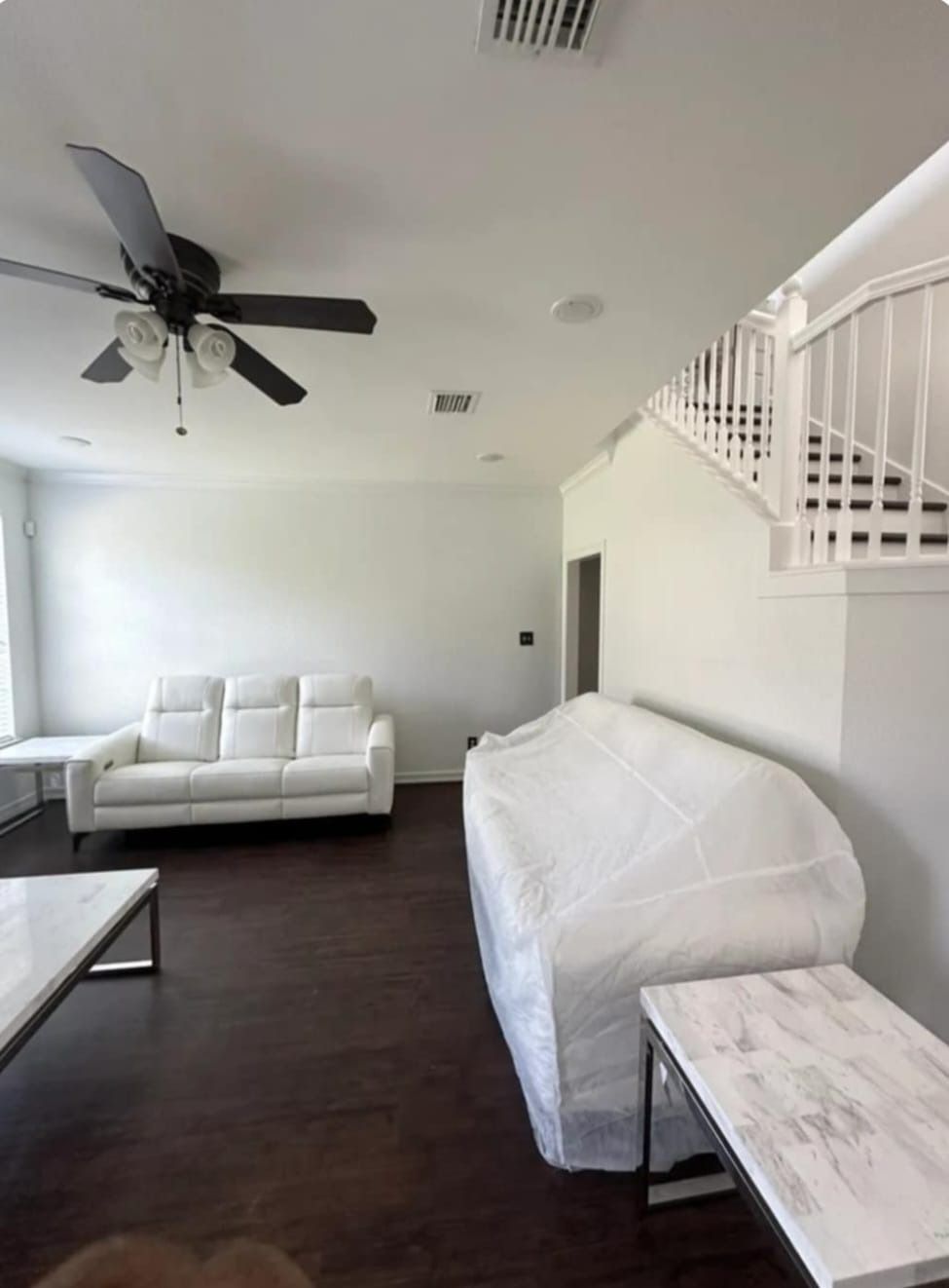 Living room with white sofa, stairwell, and plastic-covered furniture. Dark wood floors and white walls.