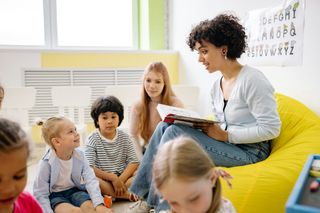 Teacher reading to children in a bright classroom, sitting on a yellow beanbag with books open.