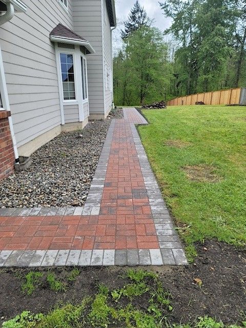Brick pathway alongside a light-colored house, leading to a grassy yard.