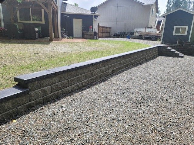 A retaining wall made of gray blocks with dark capstones, next to gravel.