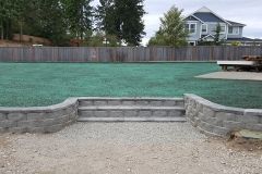 Gray stone steps lead to artificial turf lawn; fenced yard with a house in background.