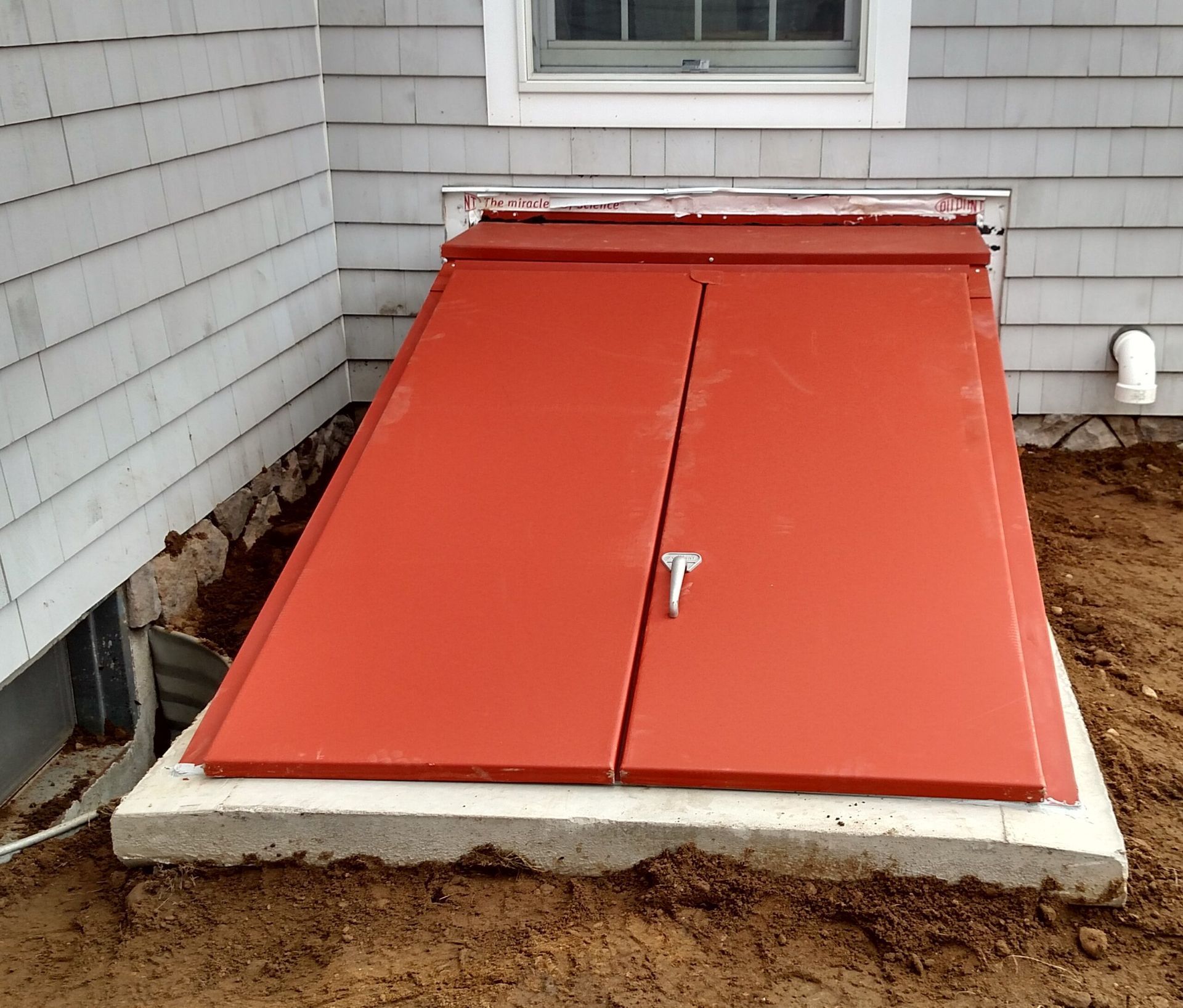 Red, slanted basement door in a concrete frame against a gray clapboard house, set in dirt.