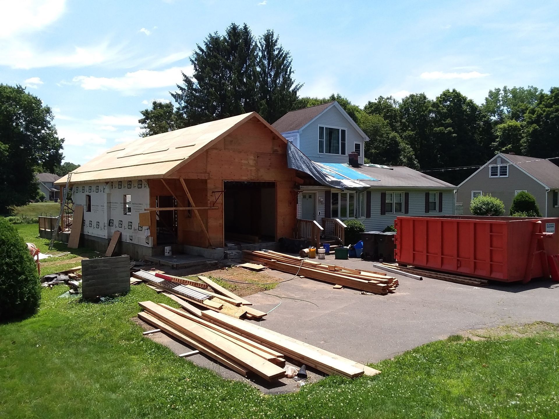 Garage under construction next to a house with blue tarp on roof, lumber scattered on driveway, sunny day.
