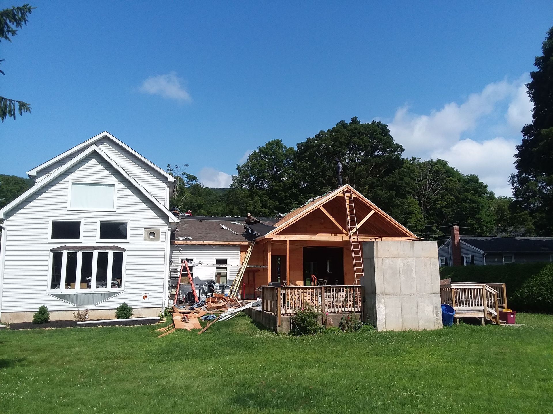 House undergoing construction on a sunny day. New wooden structure is being built next to a white building.