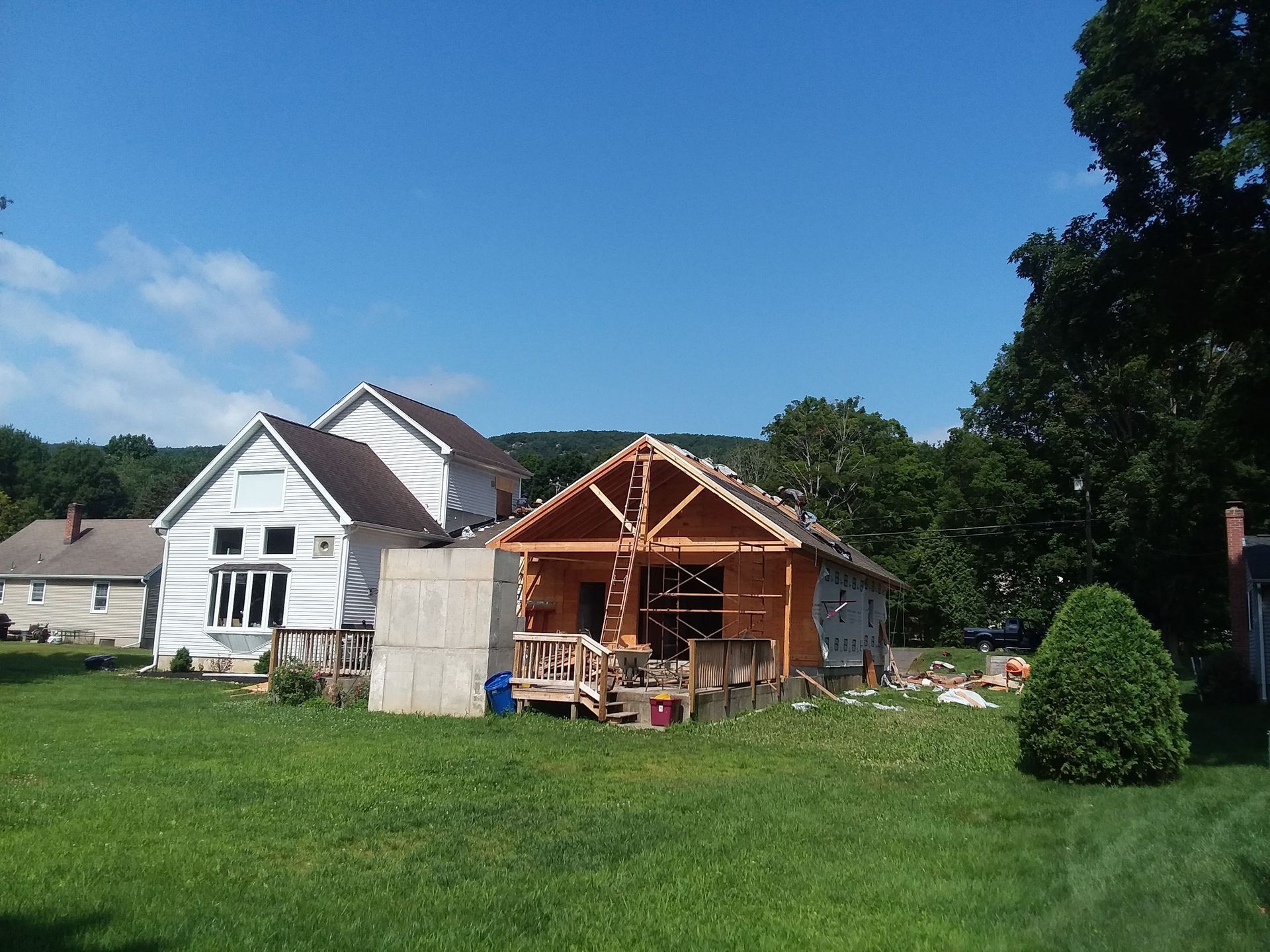 House under construction, frame visible. Adjacent to a white house, on green lawn, blue sky.
