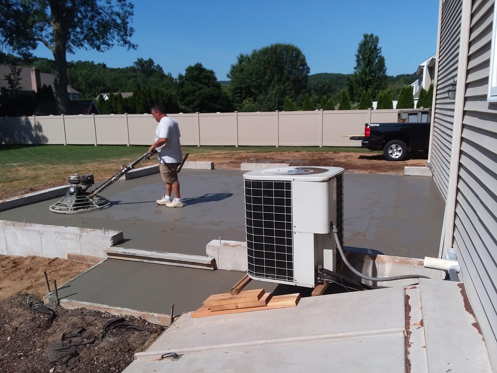 Man smoothing wet concrete with a power trowel on a patio next to a house with an air conditioning unit.