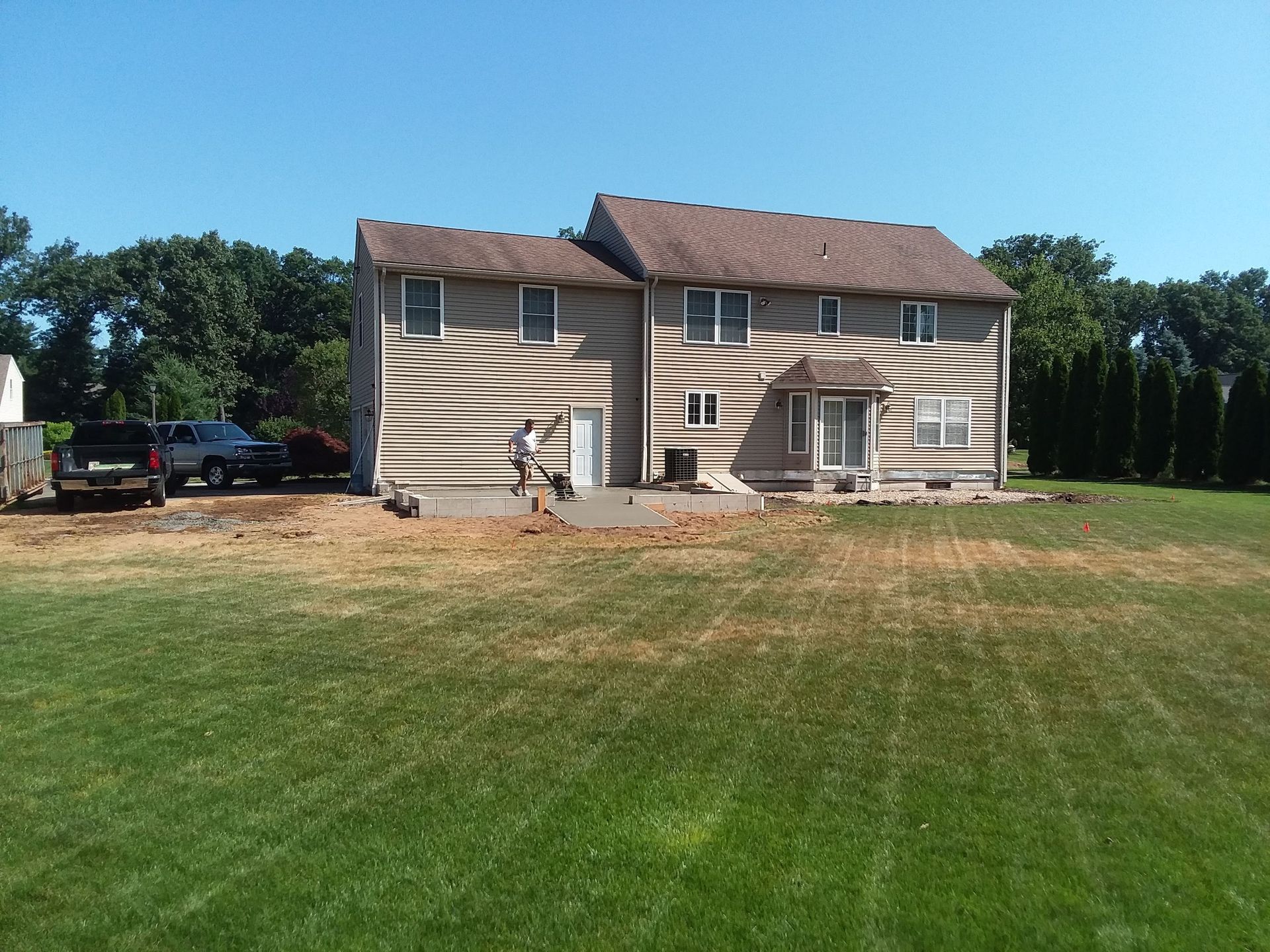 Back of a two-story house with siding, surrounded by a lawn and trees. Man stands near concrete work.