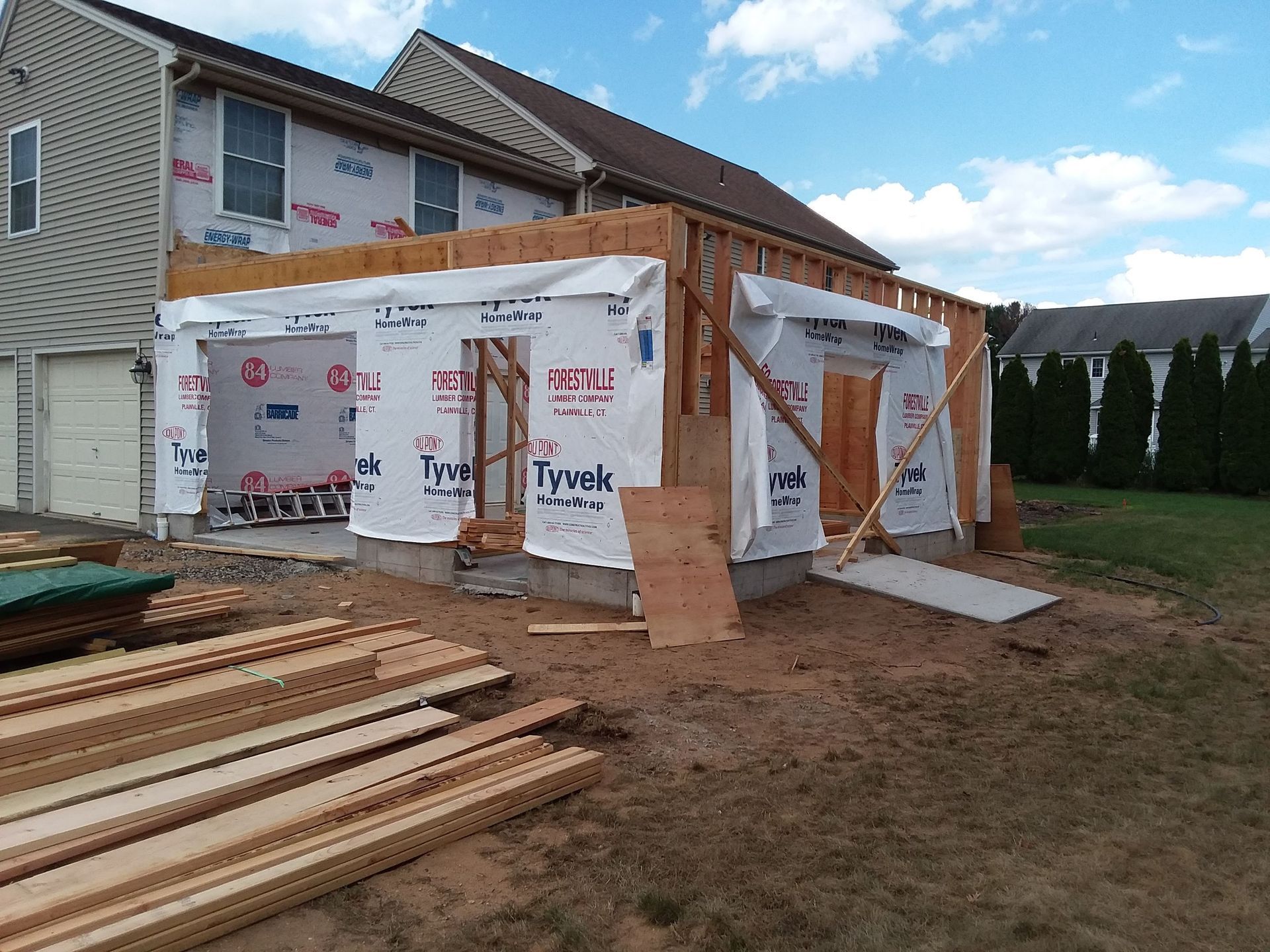 House undergoing construction, framed addition with plywood, wrapped in Tyvek. Wood beams and materials on the ground.
