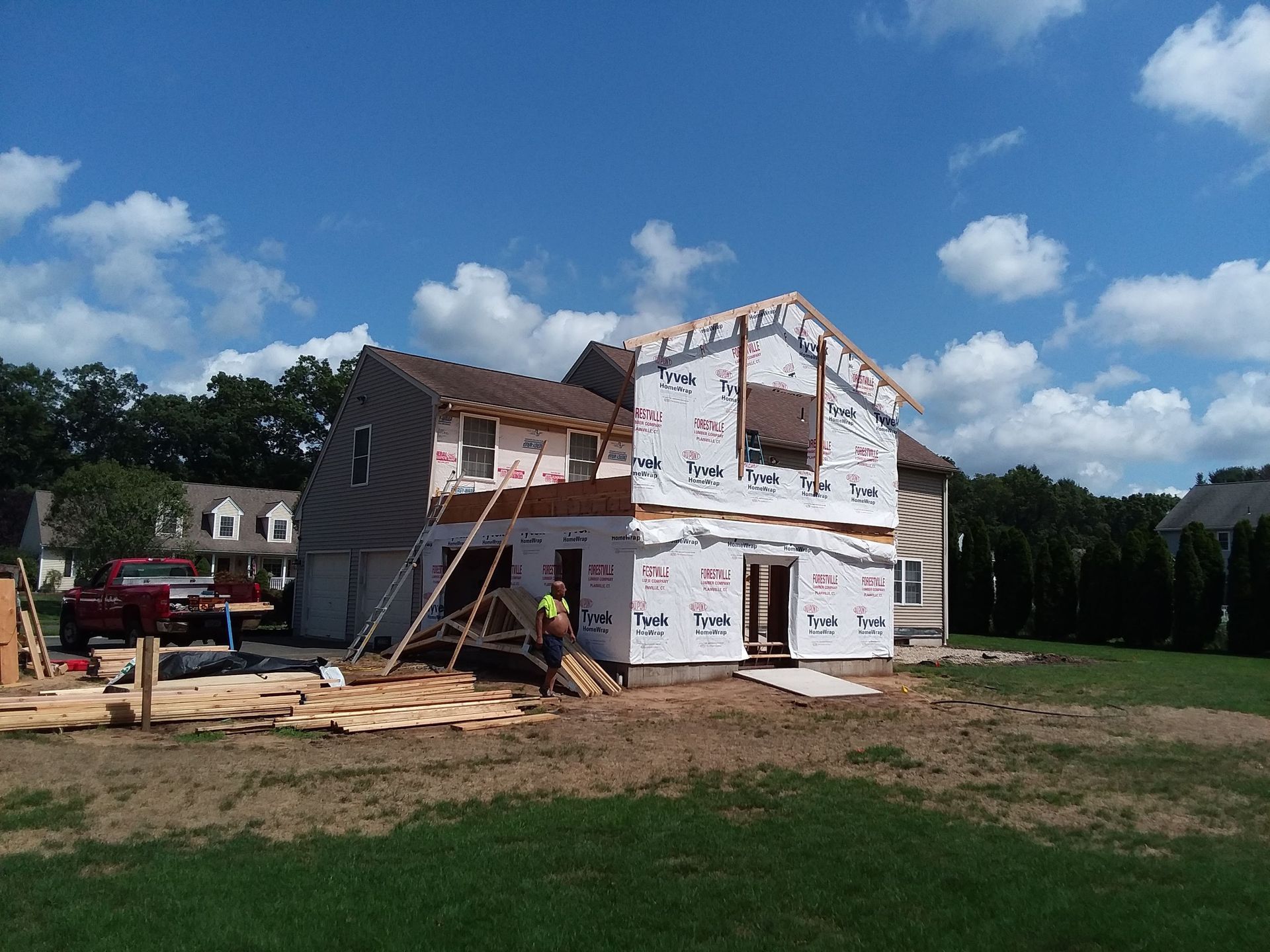 Two-story house under construction with worker, blue sky, and surrounding trees.