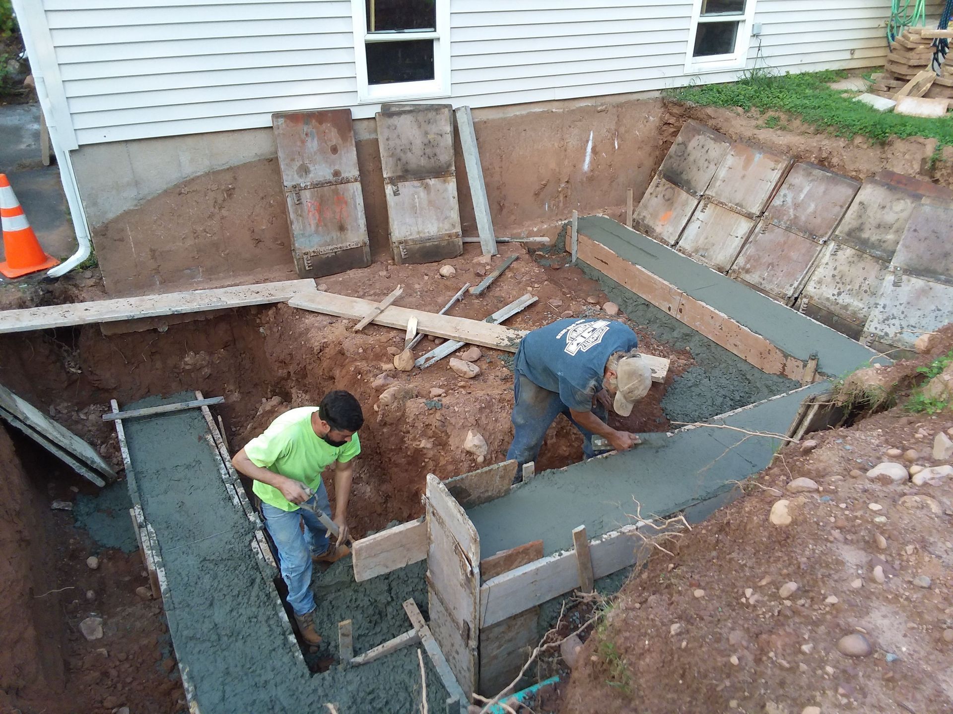 Two construction workers pouring concrete into forms for a foundation, near a house.