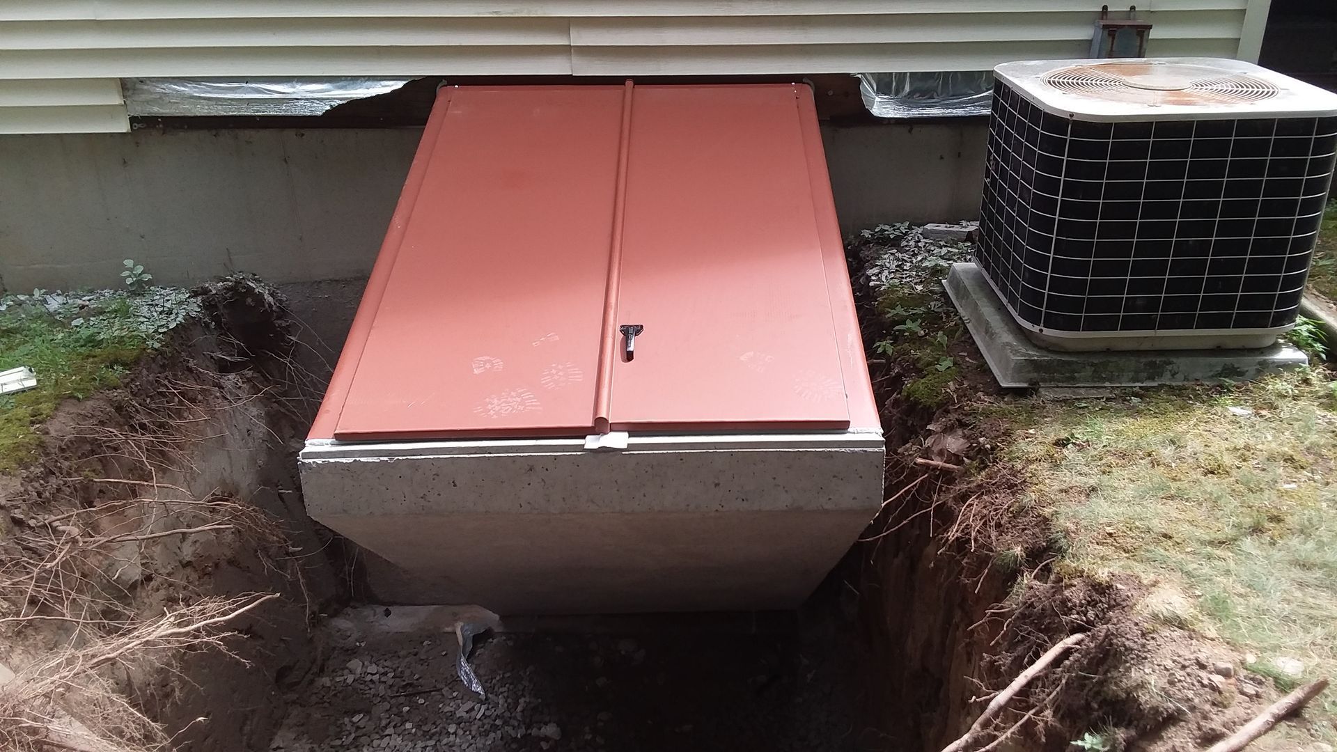 Red basement doors set in concrete, accessible via a trench; next to an air conditioner.