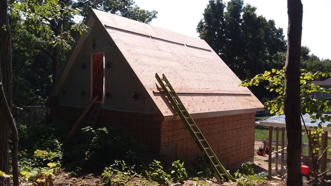 A-frame shed under construction, brown wood exterior, with a ladder leaning against it.