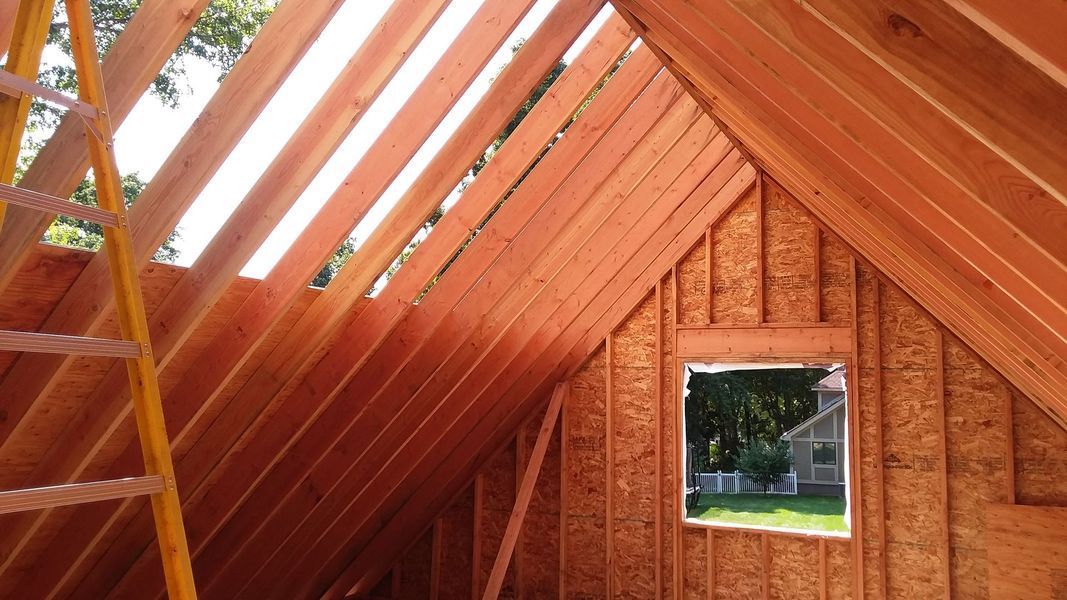 Interior of a wood-framed building under construction with a window and ladder. Sunlight streams through the roof.