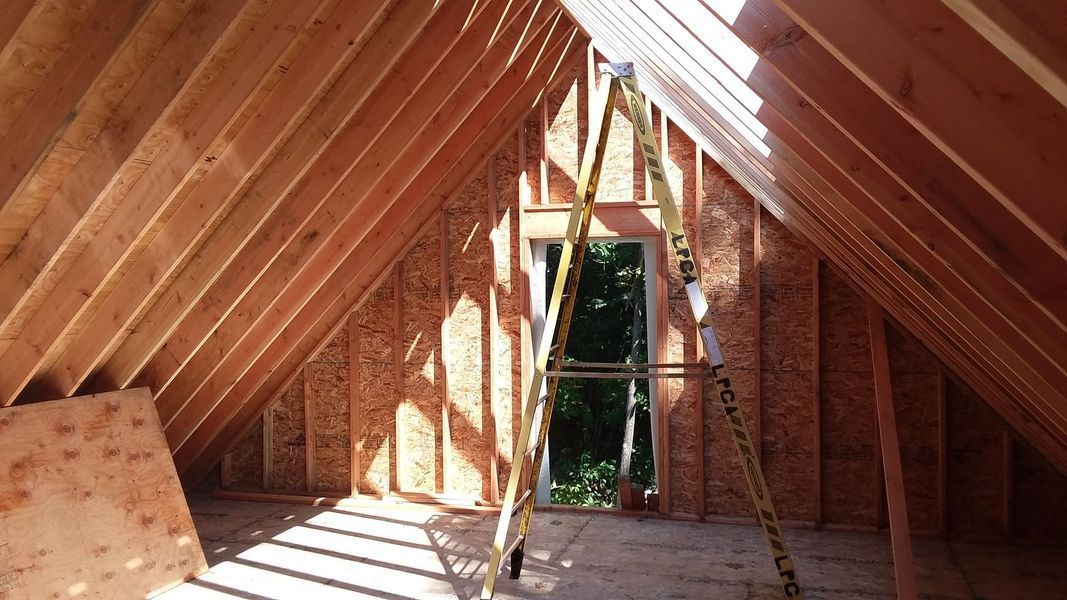 Interior of a wood-framed attic with slanted walls, a window, and a ladder inside.