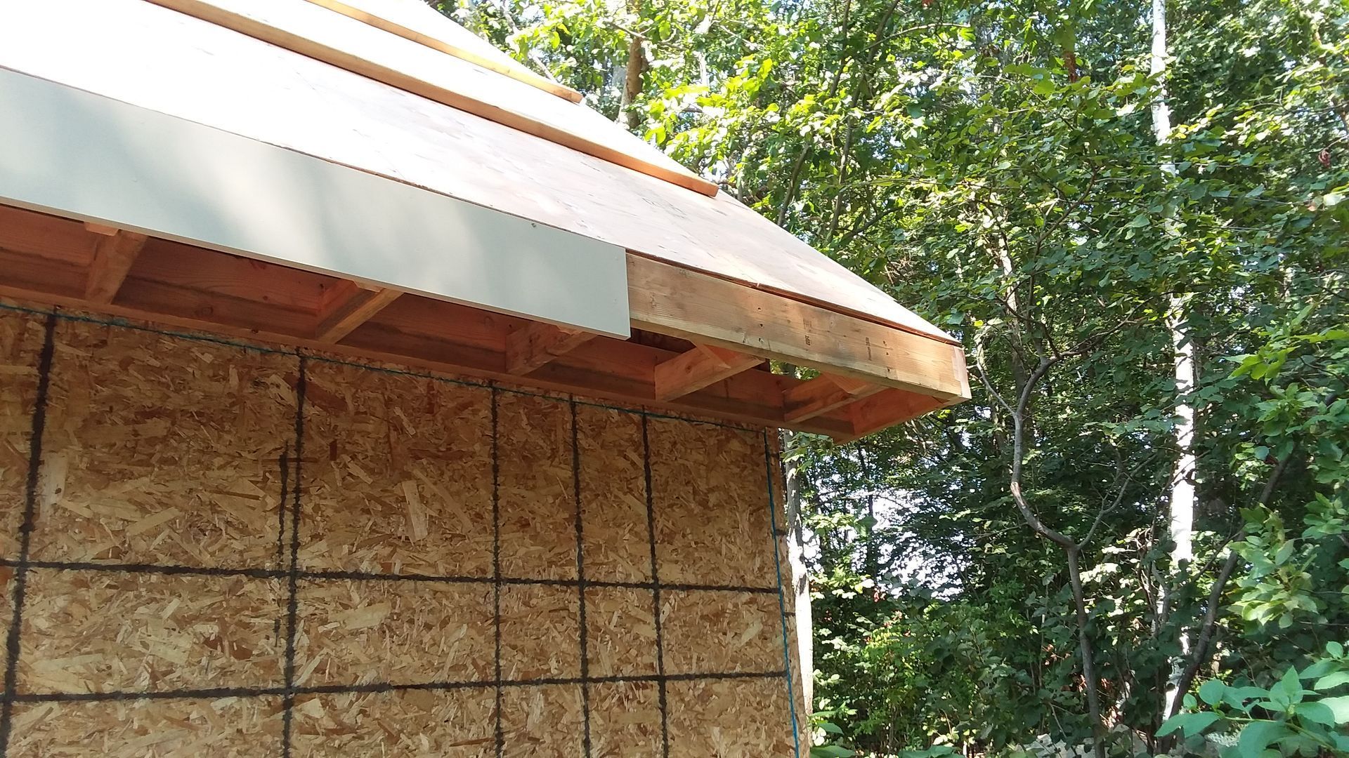A partially constructed wooden structure with an exposed roof, against a backdrop of green trees.